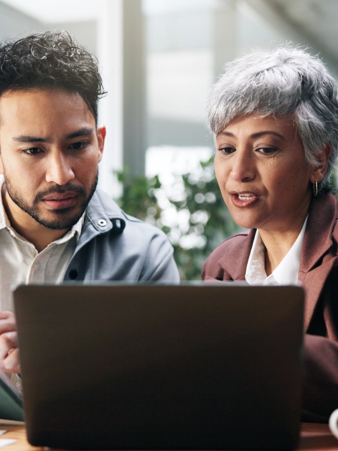 Two people sit together and look intently at a laptop screen, appearing focused and engaged in discussion in a modern indoor setting.