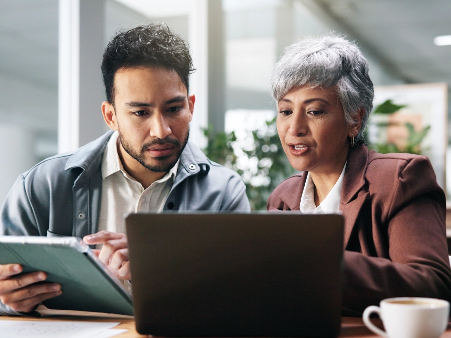 Two people sit at a desk, looking at a laptop together. One holds a tablet while the other gestures toward the laptop screen, appearing focused on their discussion.