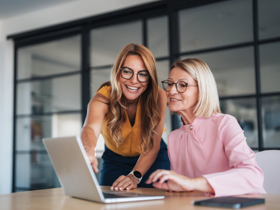 Two women wearing glasses work together at a desk, looking at a laptop screen and smiling in a modern office setting.