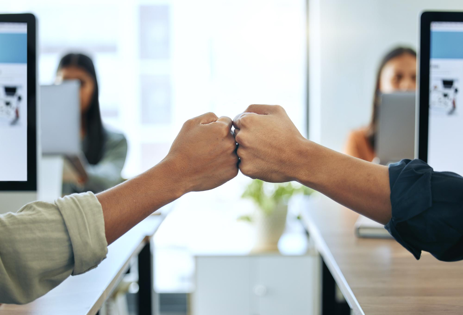 Two people fist bump across a desk in an office, while two others work on computers in the background.