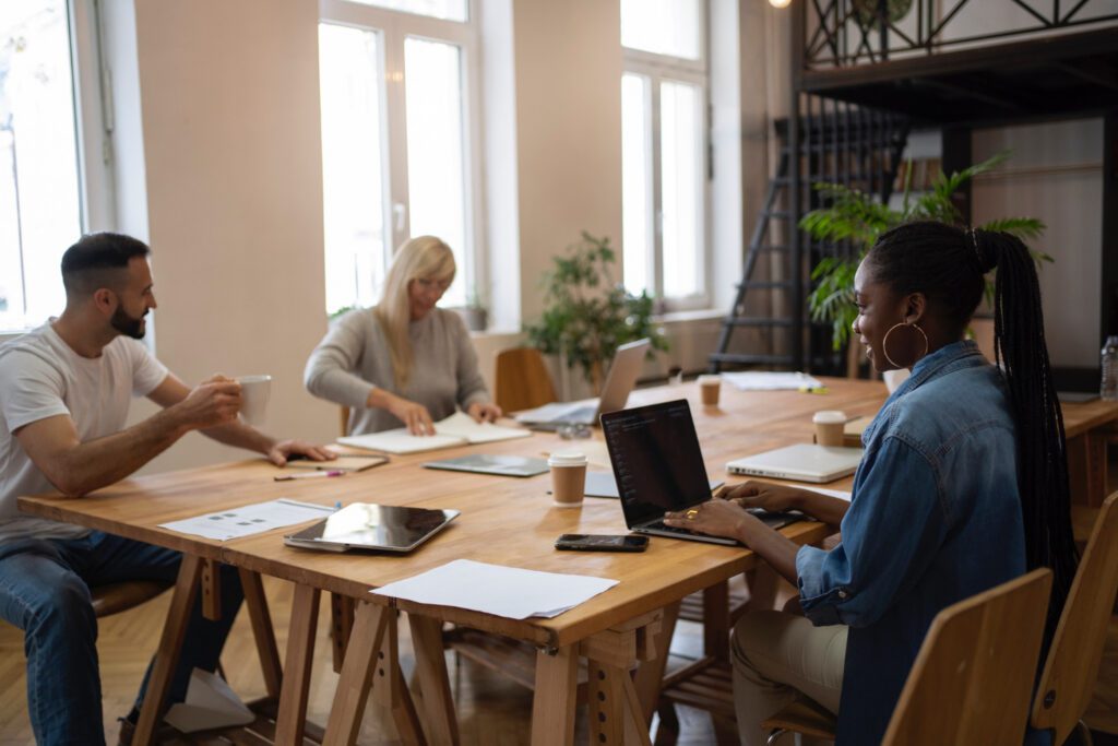 Three people are working at a wooden table in a bright office, collaborating on laptops and papers as they discuss strategies for operational enablement over coffee.