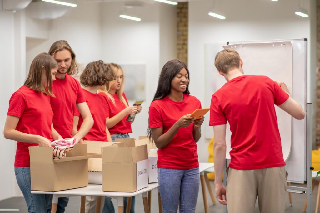 A group of people in red shirts are packing boxes and writing on a whiteboard in a well-lit room, working together on a project or volunteer activity as part of a training delivery session.