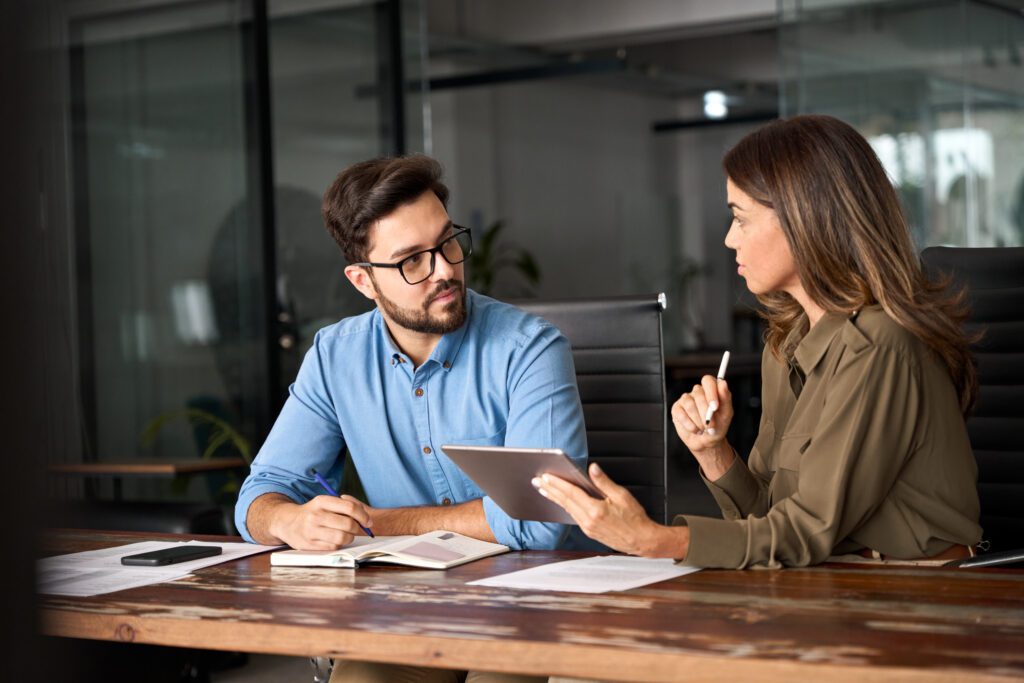 Two people sit at a desk in an office, engaged in discussion about instructional design. One holds a tablet while the other takes notes in a notebook.