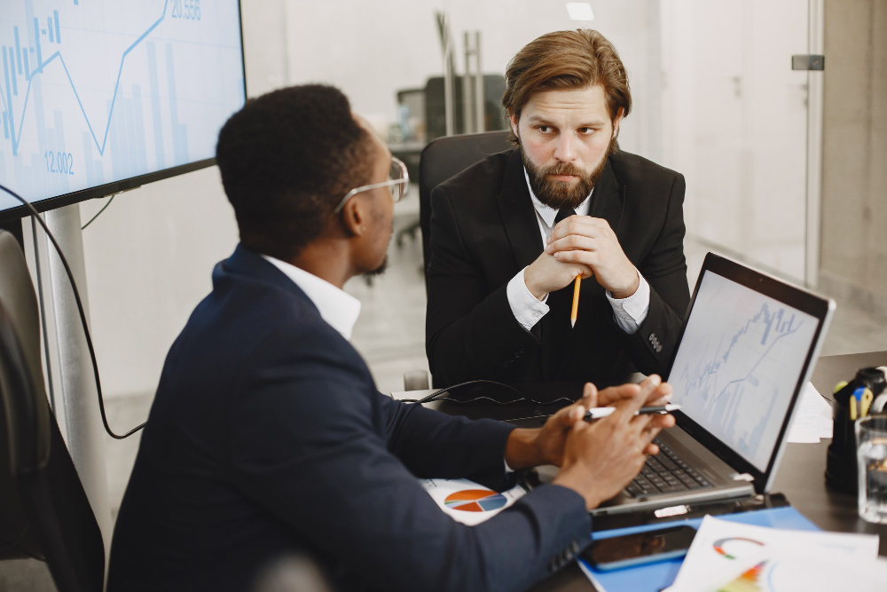 Two men in business suits discuss L&D consulting partners at a desk with a laptop, charts, and a large screen displaying a graph in an office setting, collaborating closely as L&D partners to review an evaluation guide.